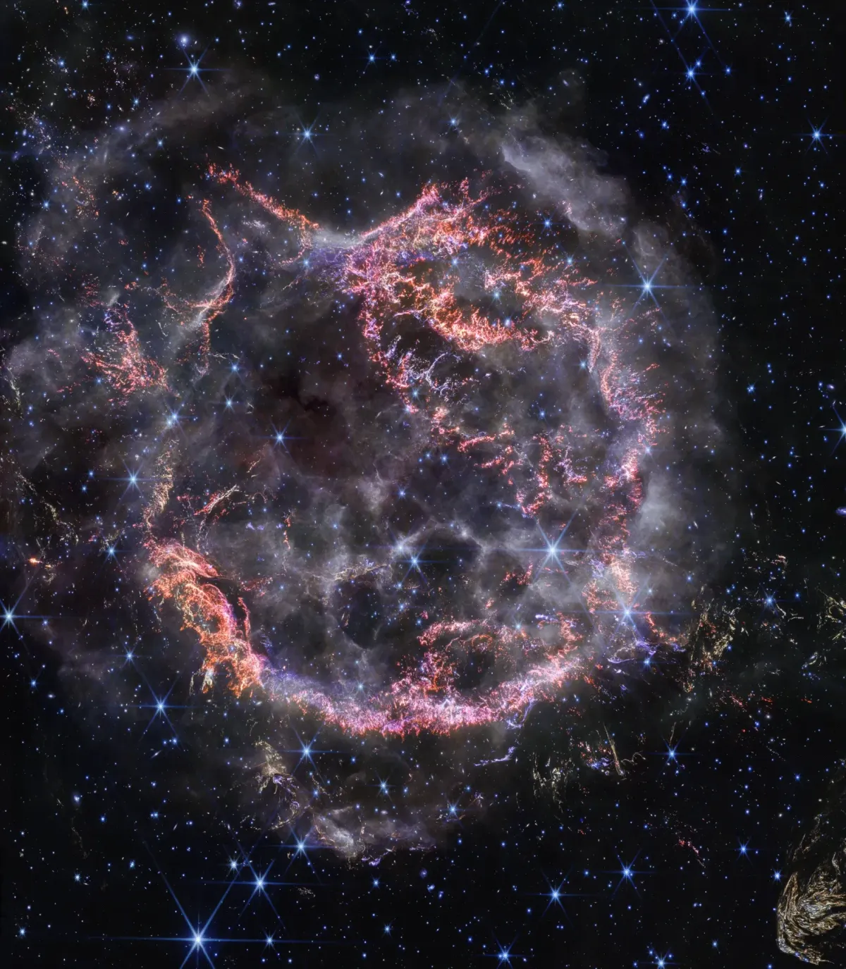 Pink and orange filaments form an expanding shell in the Cassiopeia A supernova remnant, surrounded by blue-white stars in Webb’s near-infrared view.