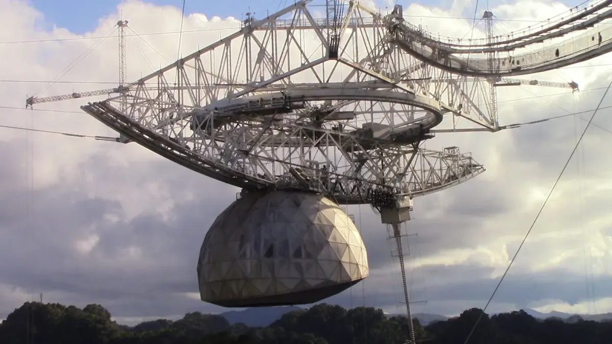 A large suspended receiver platform and rounded dome hang from cables above the Arecibo radio telescope.
