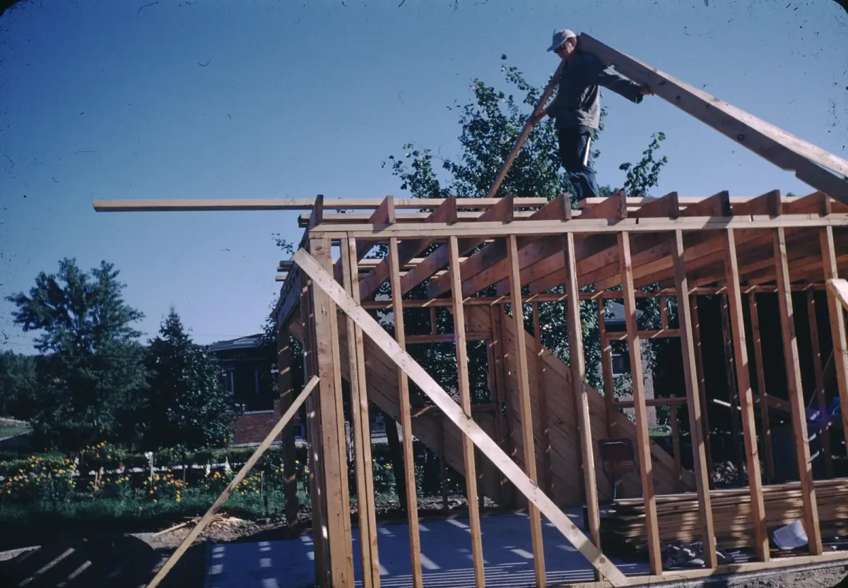 A builder carrying a beam above the exposed timber frame of a house