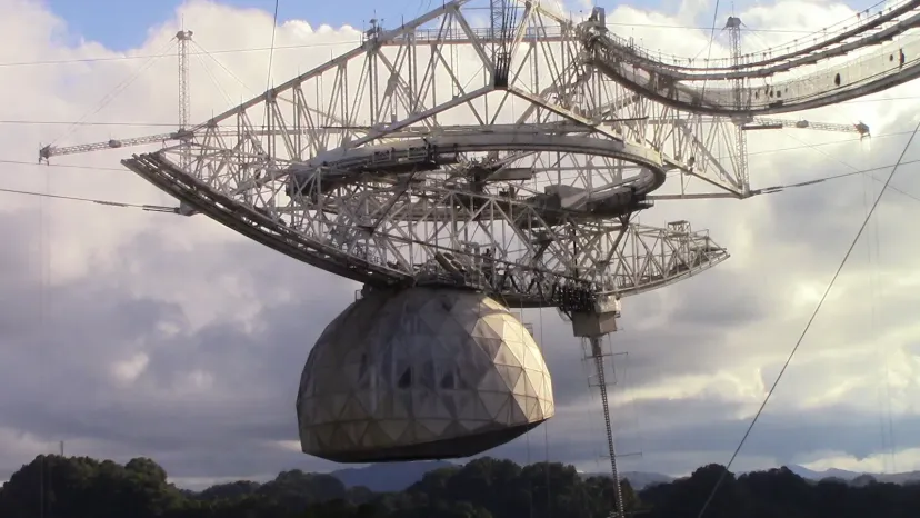 A large suspended receiver platform and rounded dome hang from cables above the Arecibo radio telescope.