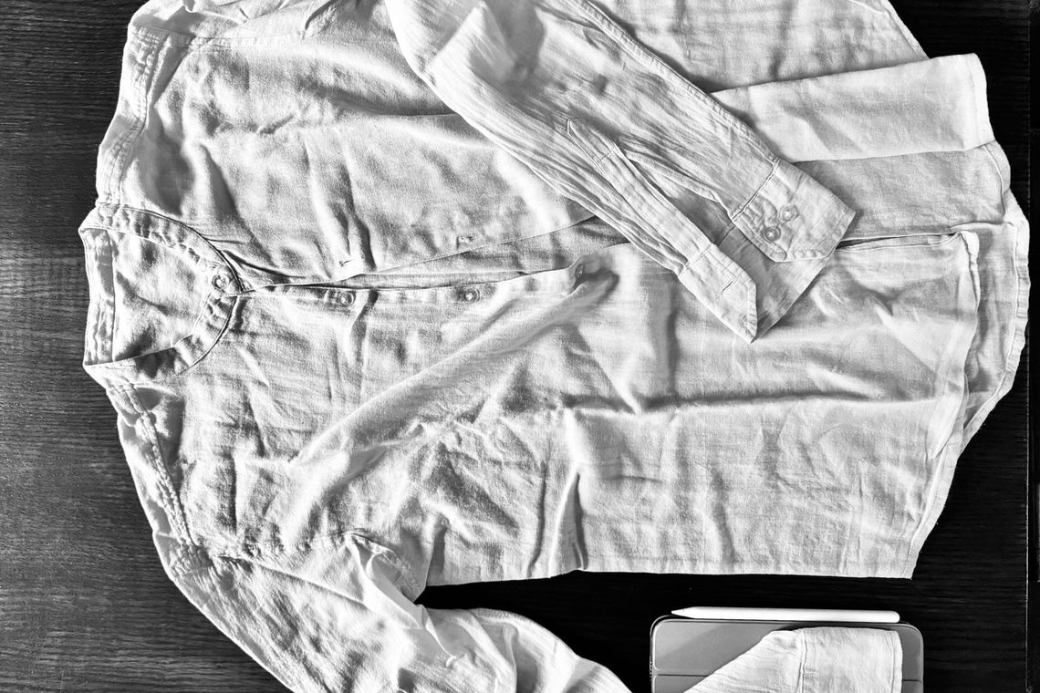 Chemise froissée en lin blanc posée sur une surface en bois sombre avec un iPad mini et son Apple Pencil dans le coin.
