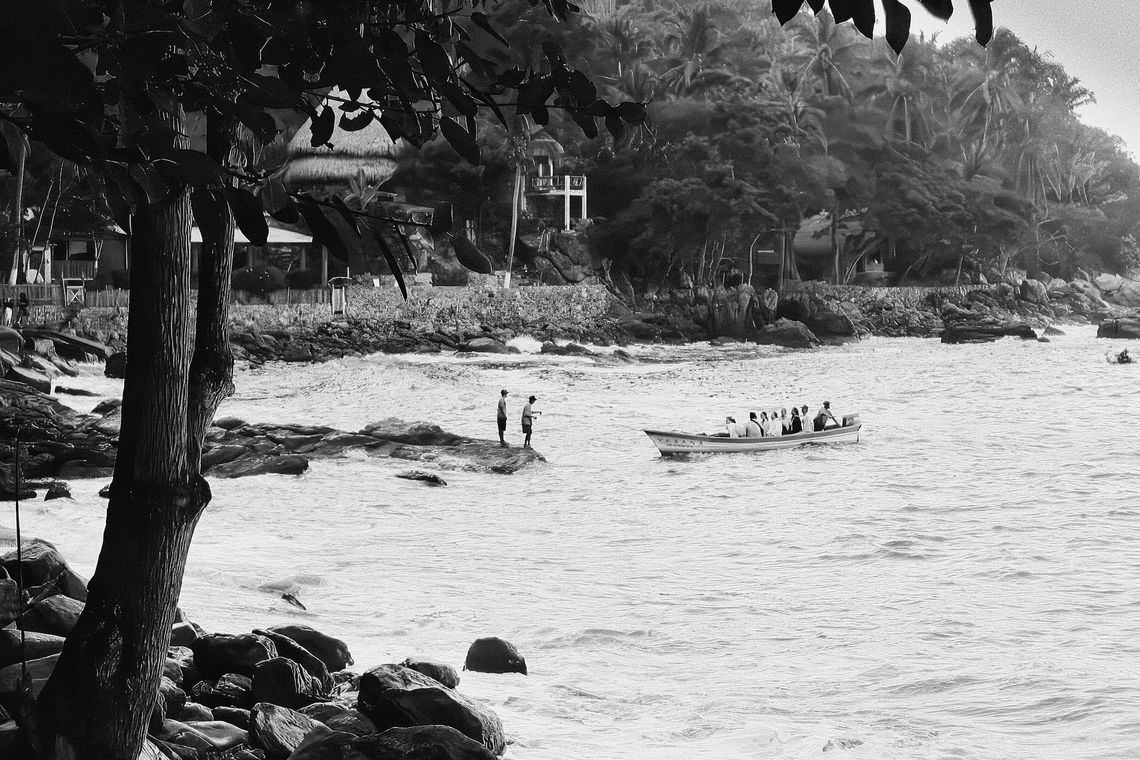 Bateau avec des passagers près d'un rivage rocheux, deux personnes sur un rocher, mer houleuse. Vue de plage, arbres et maisons en fond. Noir et blanc.