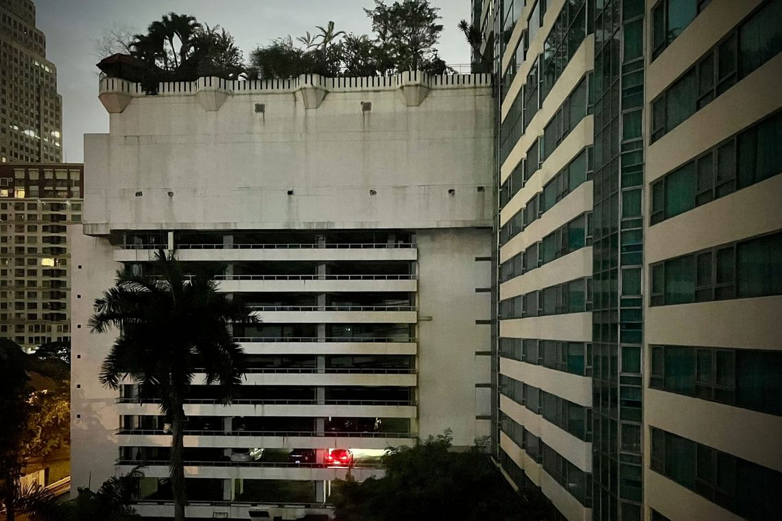 Bâtiment moderne en béton avec arbres sur le toit et un palmier solitaire, entouré d'autres immeubles, lumières de la ville le soir. En bas au milieu, feux rouges arrières d'une voiture.