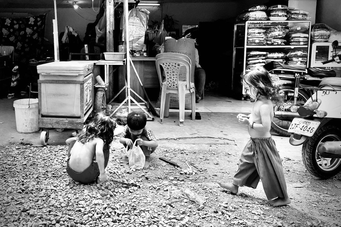 Trois enfants jouent sur un sol en gravier devant un magasin, entourés de vêtements et d'un scooter garé.