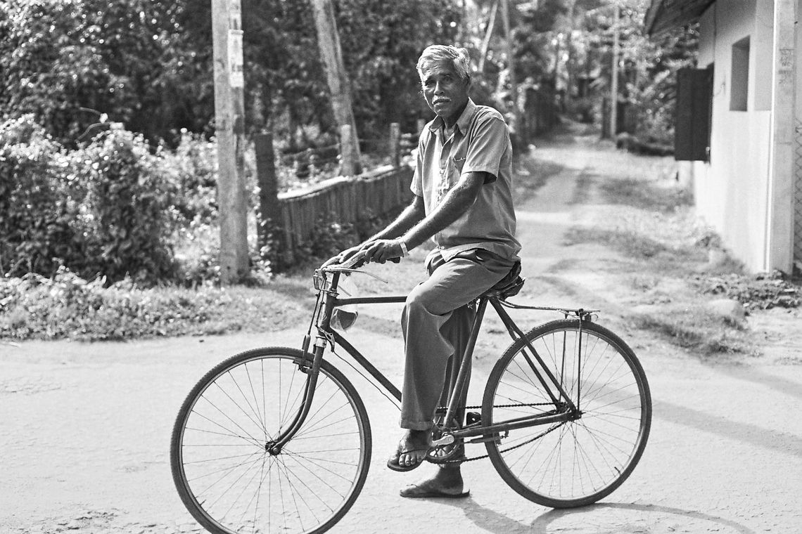 Un homme âgé (Adakkan), cheveux et moustache blancs, pose paisiblement assis sur son vélo dans une rue bordée d'arbres, vêtu simplement, avec des sandales. L'image est en noir et blanc.