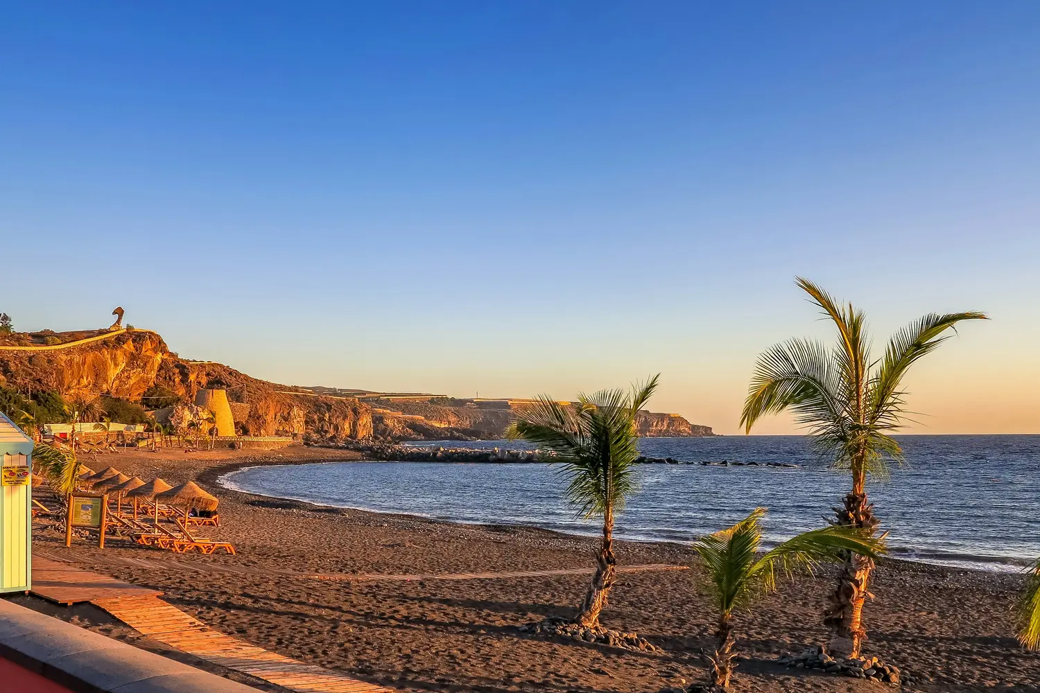 Tropical beach with palm trees, dark sand, and rocky cliffs at sunset in Spain