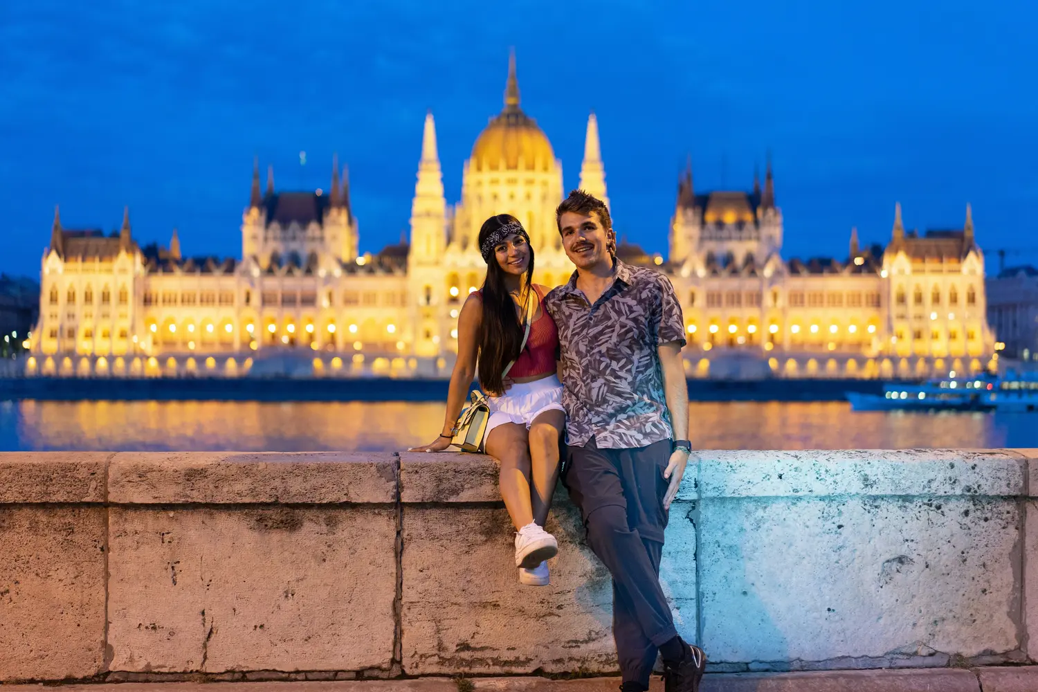 A couple posing on a riverside wall in front of the illuminated Hungarian Parliament Building in Budapest, Hungary.