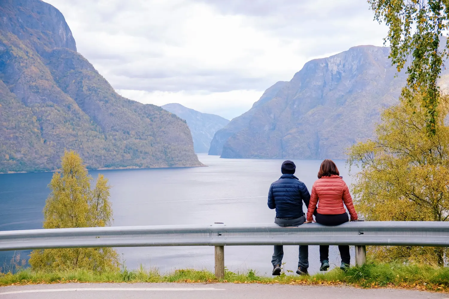 A couple sitting on a metal roadside guardrail overlooking a scenic fjord in Norway