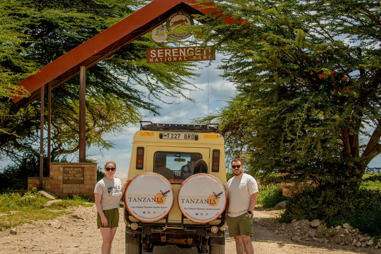 Two visitors posing in front of an official entrance gate at Serengeti National Park in Tanzania