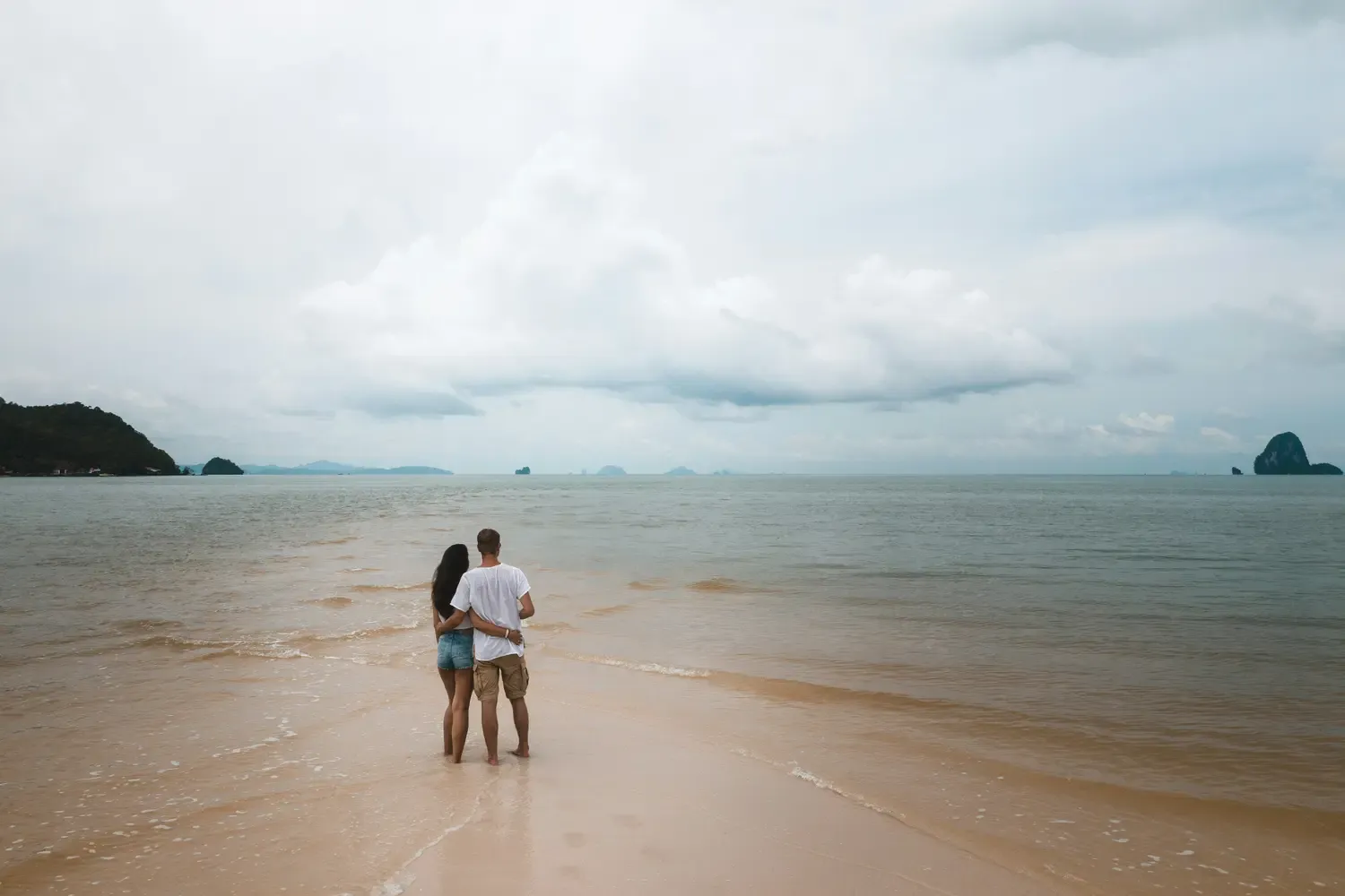 A couple standing on a sandy beach looking out at a calm sea under an overcast sky