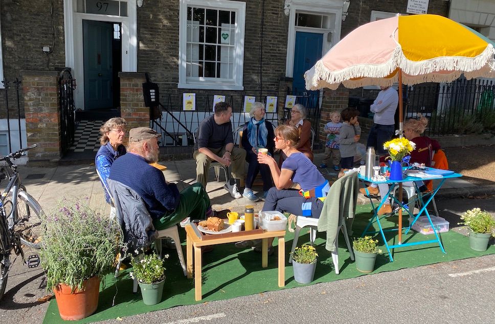 8 adults and 2 children sit and chat in a parklet. There are pot plants and a sun umbrella.