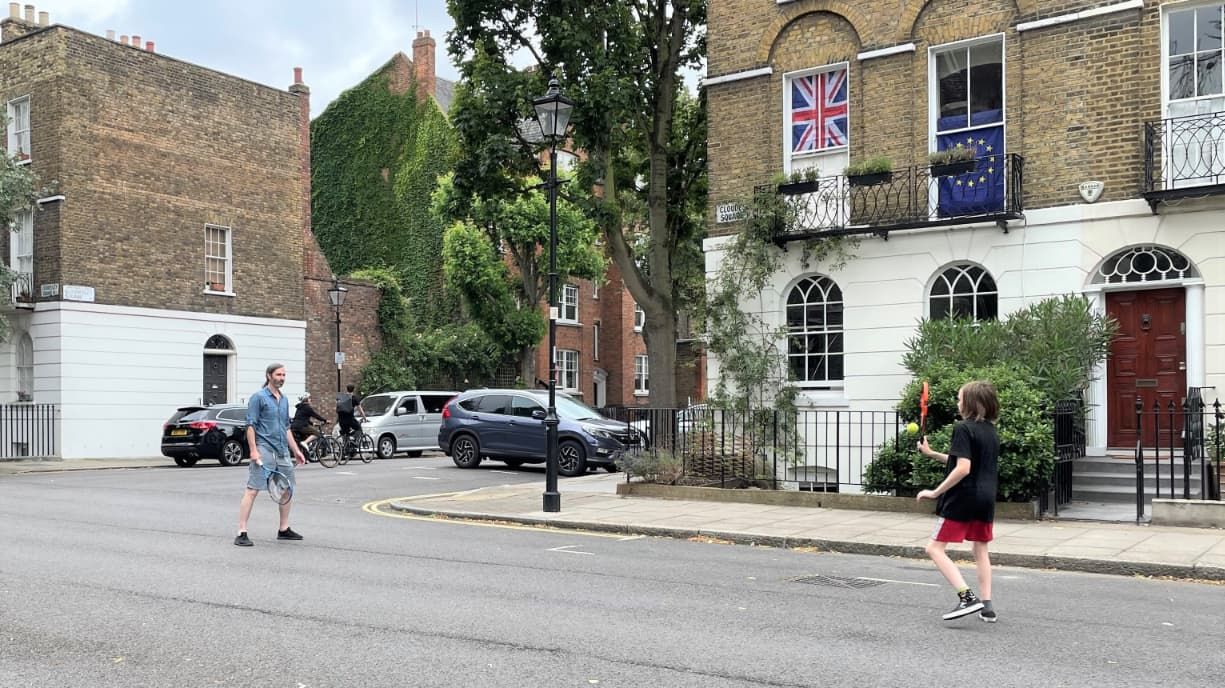 A man and a boy play tennis in the middle of a low-traffic street in a neighbourhood of terraced houses.