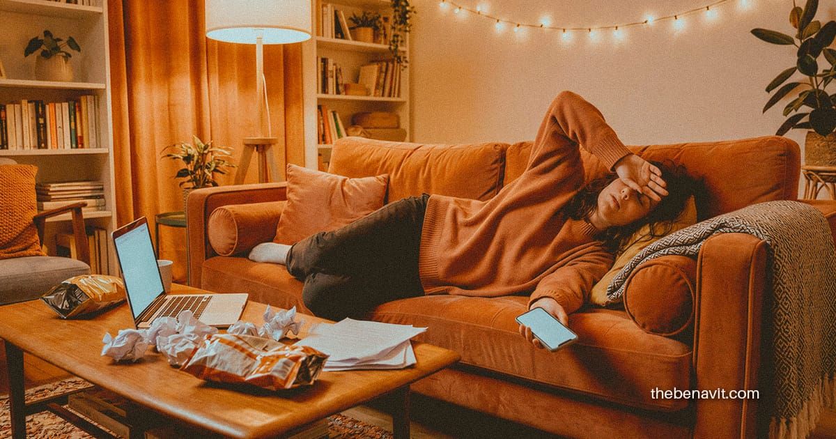 A person lying dramatically on a couch ignoring an open laptop, phone in hand, surrounded by snacks and crumpled paper.