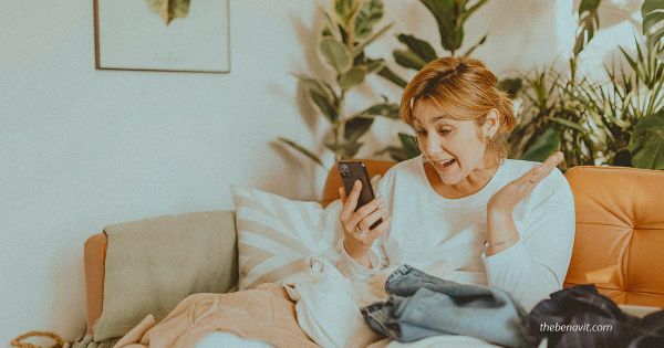 A person sitting on a couch surrounded by shopping bags, smiling at their phone after a purchase.