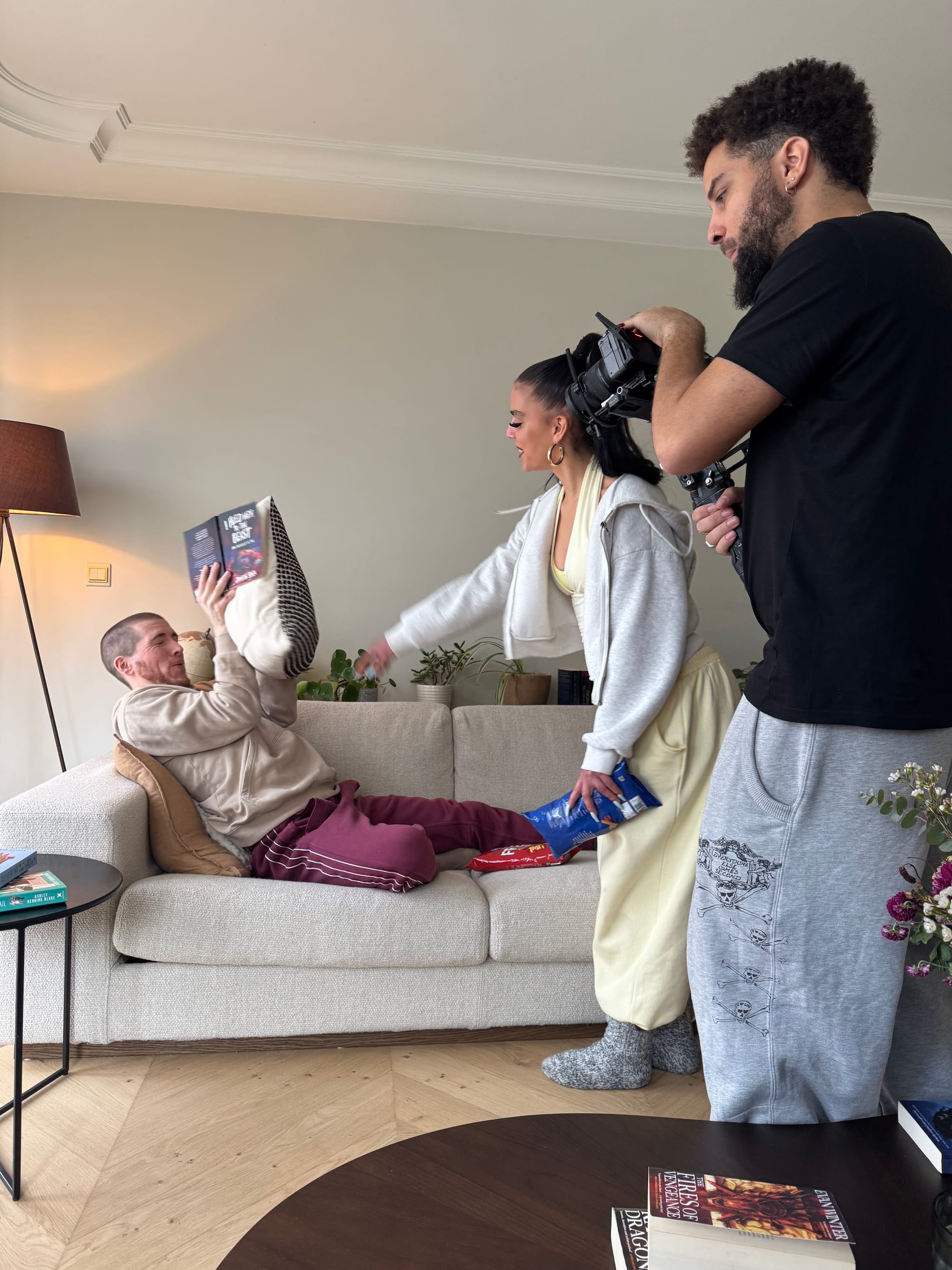 Slim B is sitting in the couch and reading a book. Pippa throws a pillow at him. Adam is filming them.