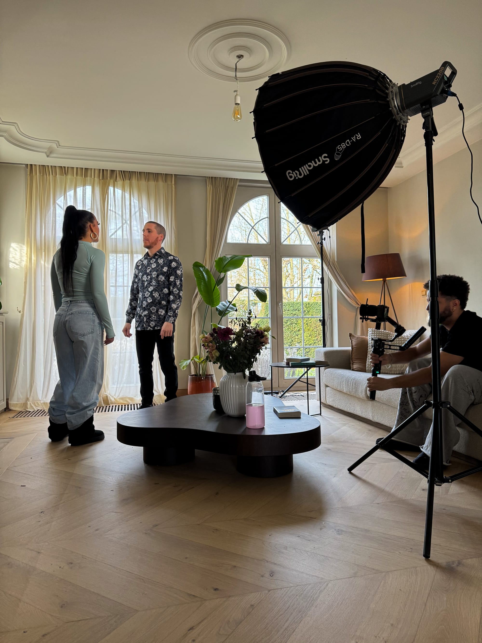 Pippa and Slim B are standing in the corner of the living room, looking at each other. Adam is sitting on the couch and filming them.