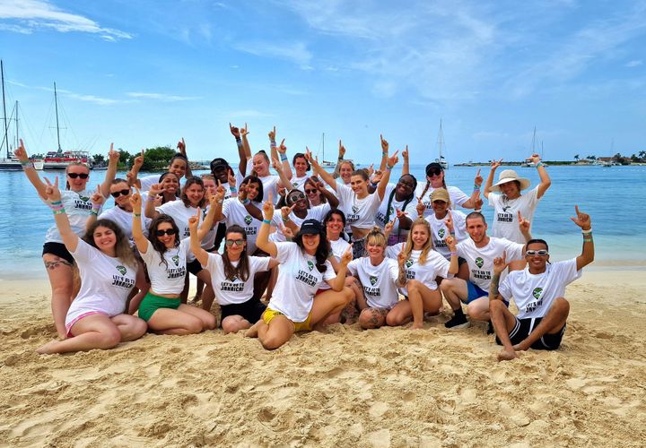 Group picture of 27 people on the beach with their back to the sea. They are pointing both their index fingers up to the sky.