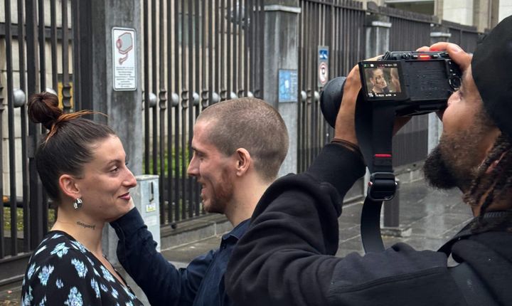 Slim B is putting his right hand on Todz cheek. They are looking at each other. Adam is to the right filming this.