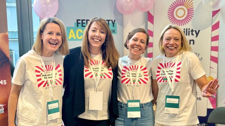 Four women with matching t-shirts smiling in front of a Fertility Action banner. 
