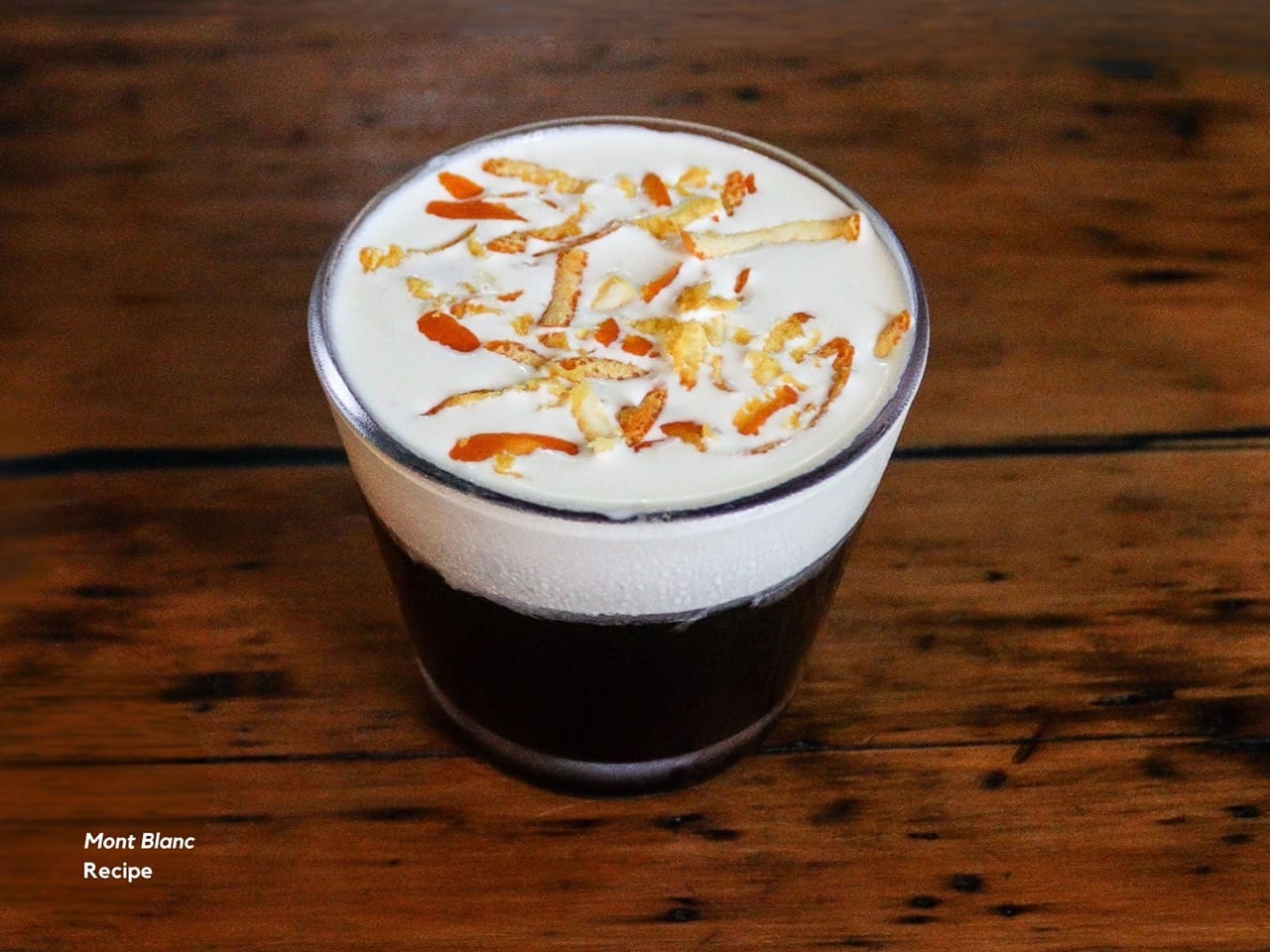 Close-up of a coldbrewed coffee topped with foam and orange zest, served on a wooden table in a minimalist café setting.