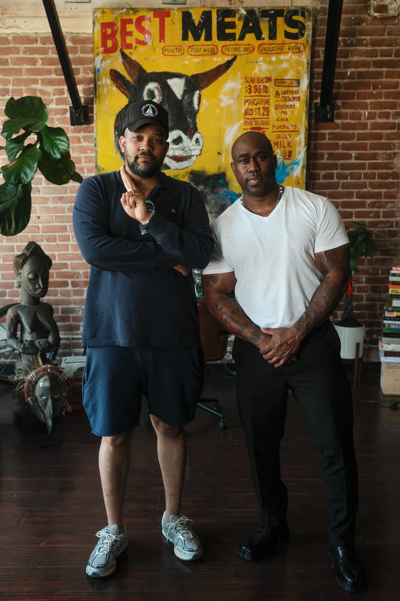 Perri Dash stands beside LA-based figurative artist Ronnie Robinson in an art-filled studio with brick walls, sculptures, and a bold yellow painting in the background.