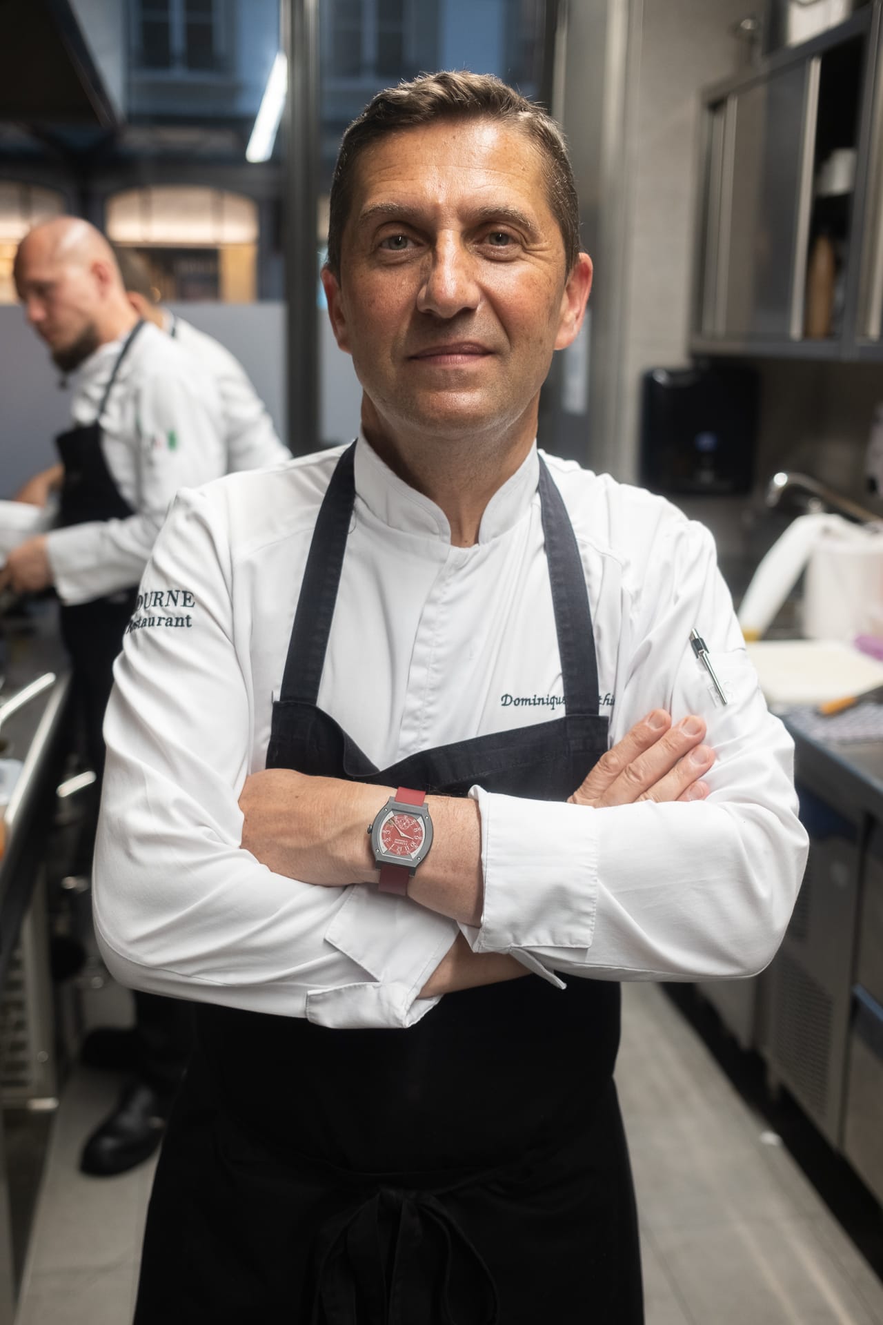 The head Chef Dominique Gauthier standing in the kitchen with folded arms, wearing his chef whites and a red F.P. Journe watch on his wrist.