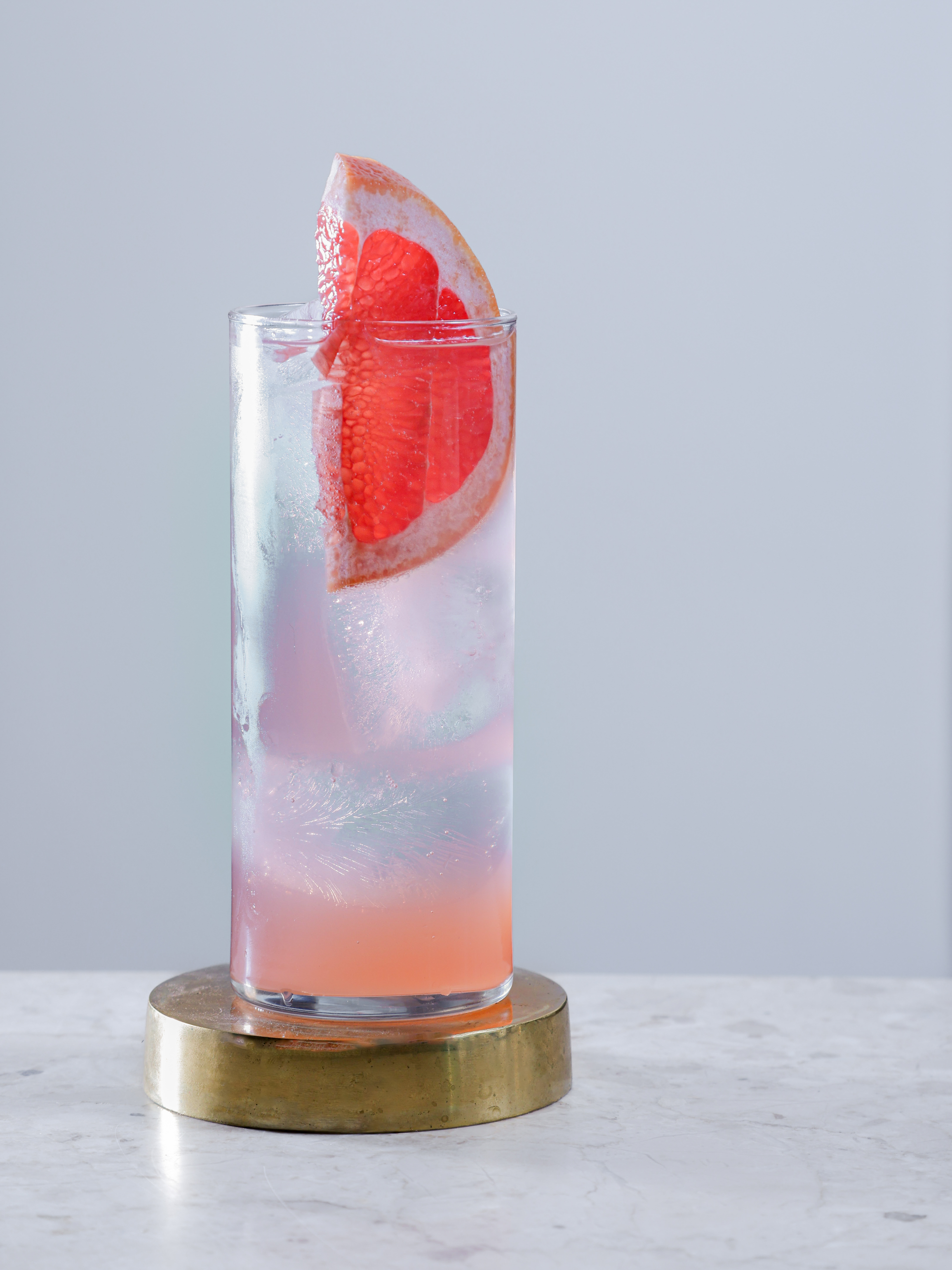Tall glass of Julia Gin cocktail with pink grapefruit garnish on a brass coaster, featuring soft pastel tones and minimalist styling on marble surface.