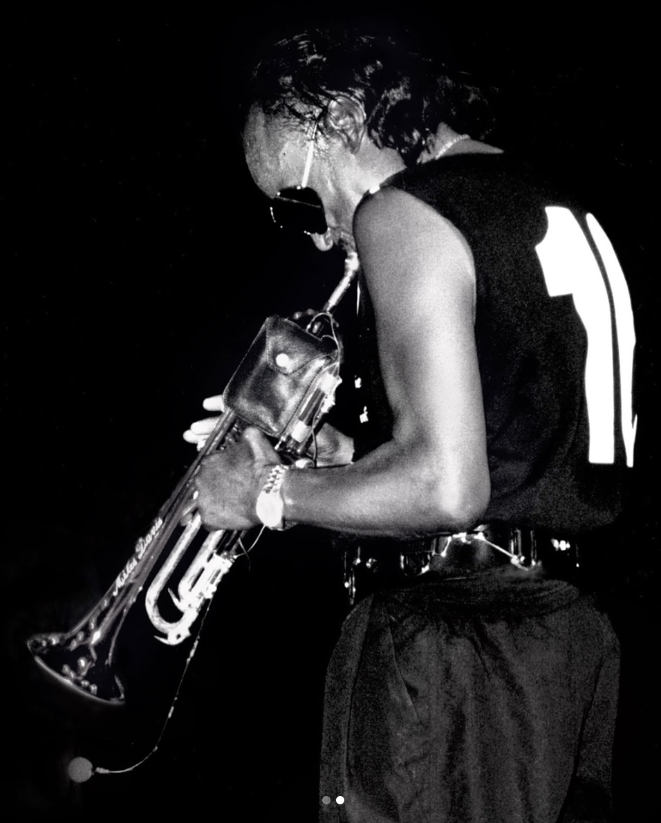 Black and white photo of Miles Davis playing trumpet on stage, wearing a wristwatch and sunglasses — a portrait of rhythm, precision, and timeless style.