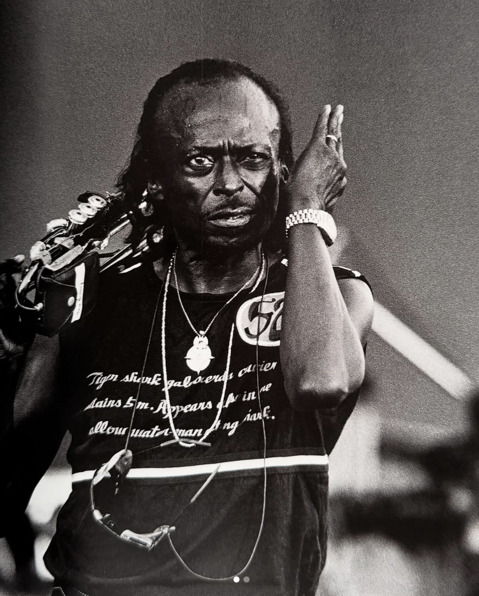 Black and white portrait of Miles Davis holding his trumpet and wearing a wristwatch and layered necklaces, captured mid-performance with expressive intensity.