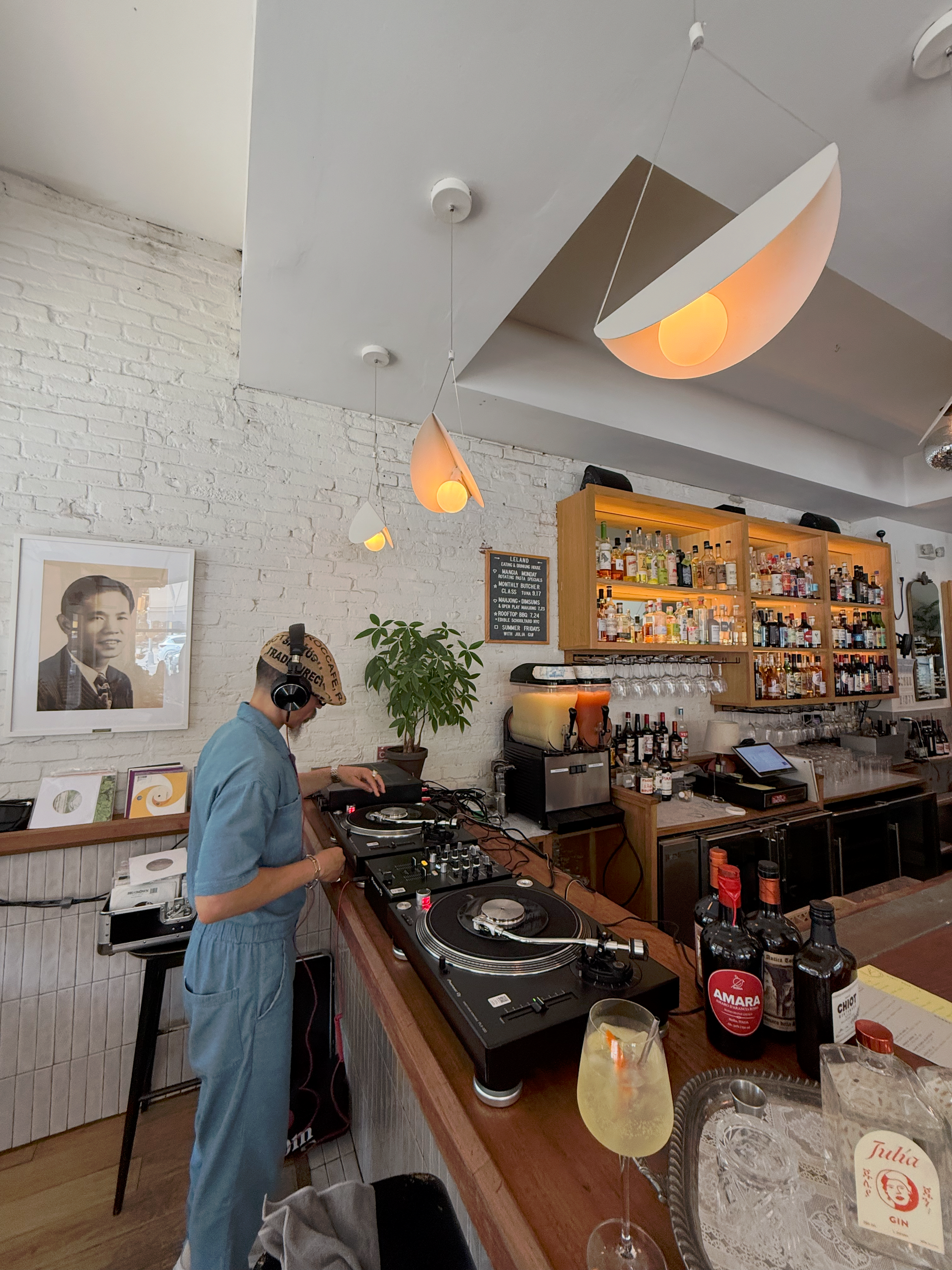 DJ in a blue jumpsuit mixing vinyl at a cozy bar with modern pendant lights, bottles of Júlia Gin and Amara on the counter, capturing a refined daytime music and cocktail scene.