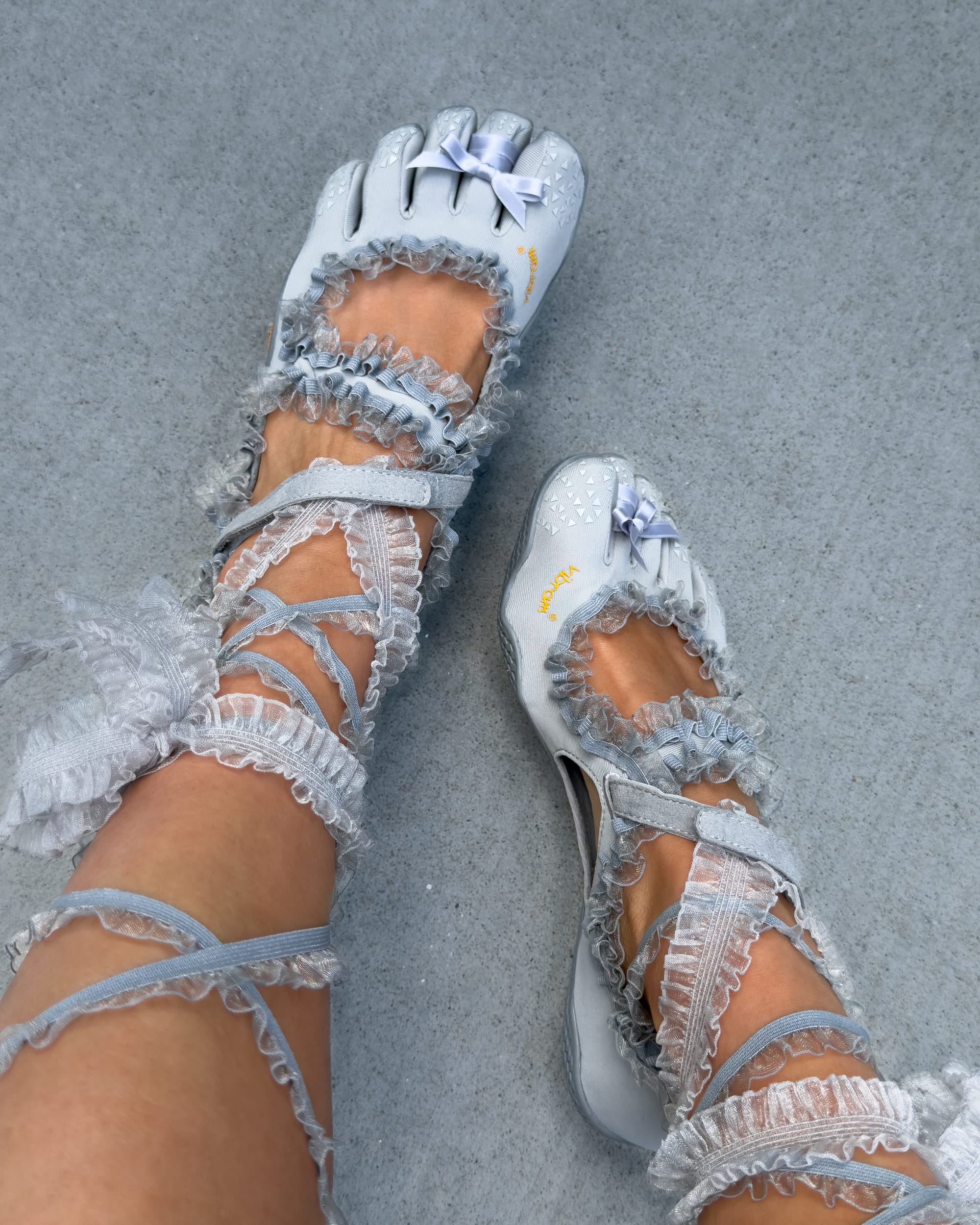 Close-up of feet wearing grey Vibram toe shoes reimagined with delicate lace ribbons and bows, merging performance footwear with romantic, avant-garde design.