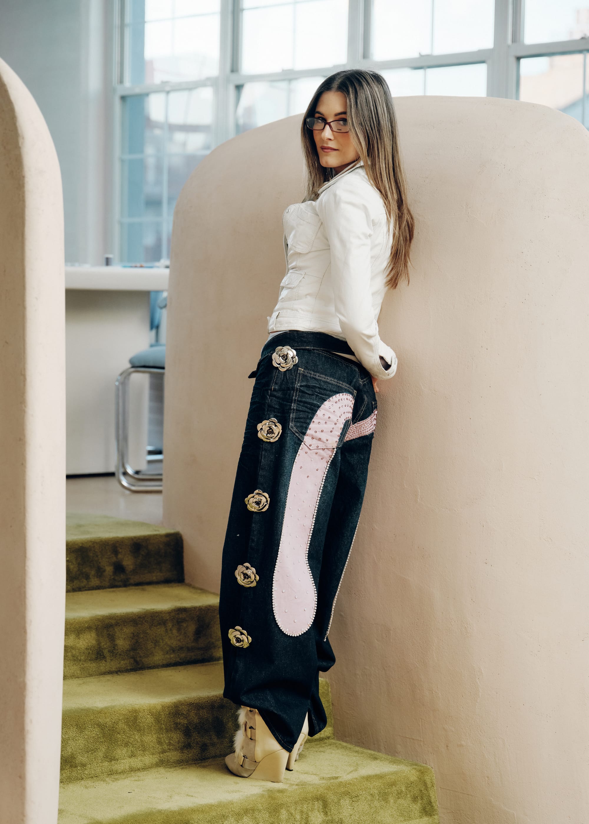 Caterina Mongillo, aka Metagirl wearing sculptural denim trousers with floral detailing and a white top, standing on carpeted stairs in a softly lit studio.