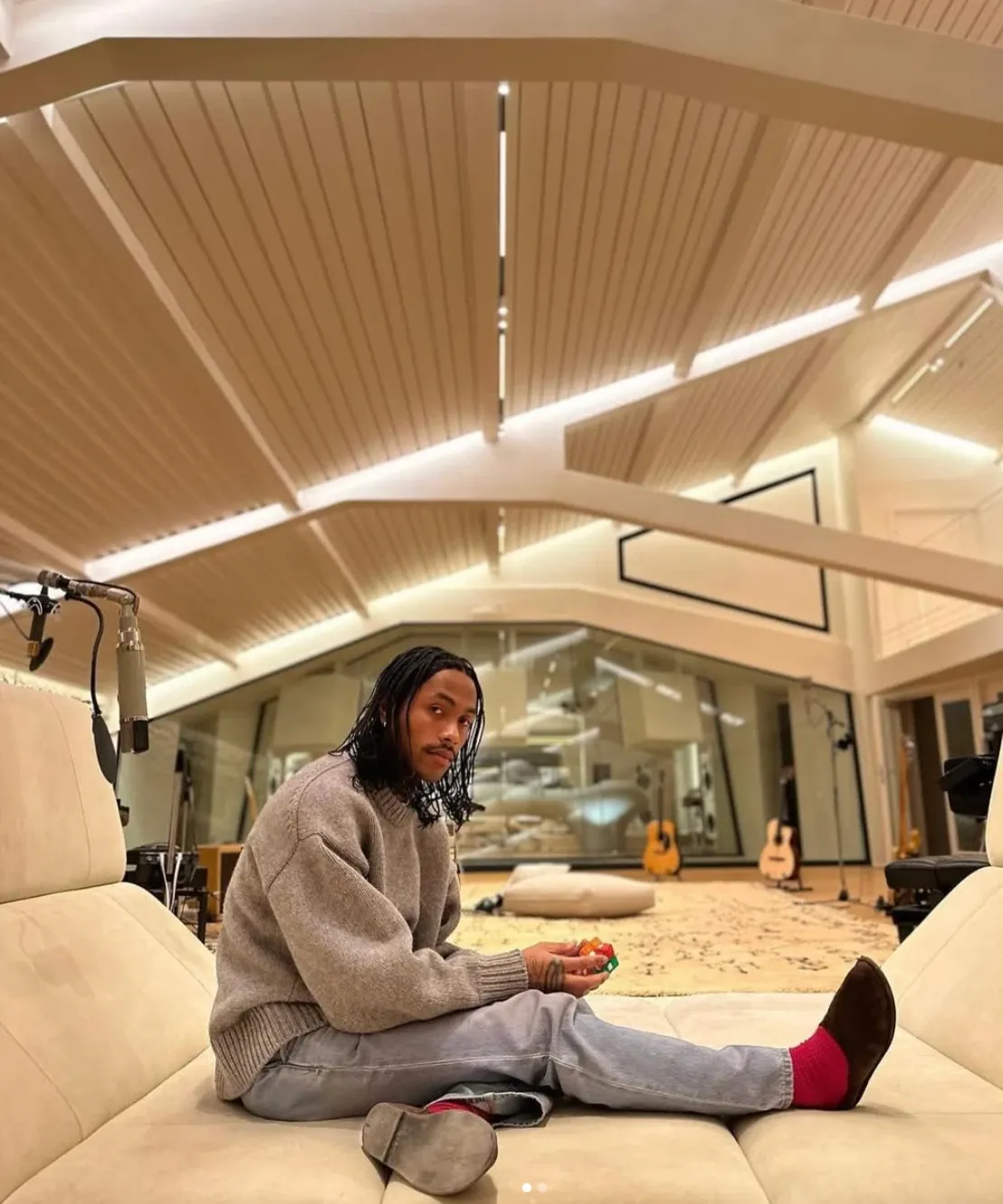 A person seated in a modern beige living room, looking into the camera on a curved Pierre Paulin sofa under ribbed ceiling lights.