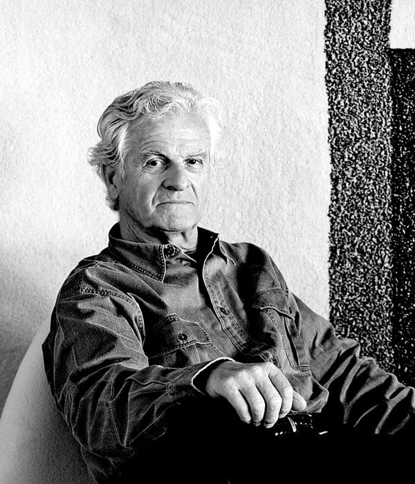 Black-and-white portrait of Pierre Paulin sitting casually in a chair, wearing a denim shirt, looking toward the camera.