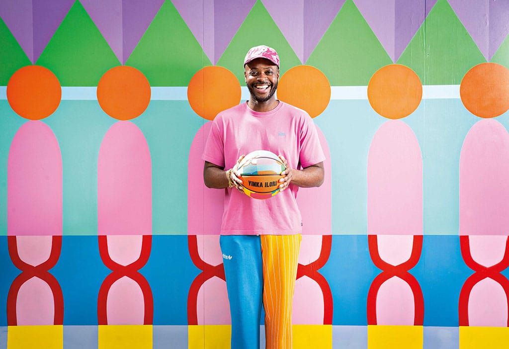 Artist standing in front of a colorful geometric mural holding a basketball painted in matching patterns, embodying play, culture, and design.