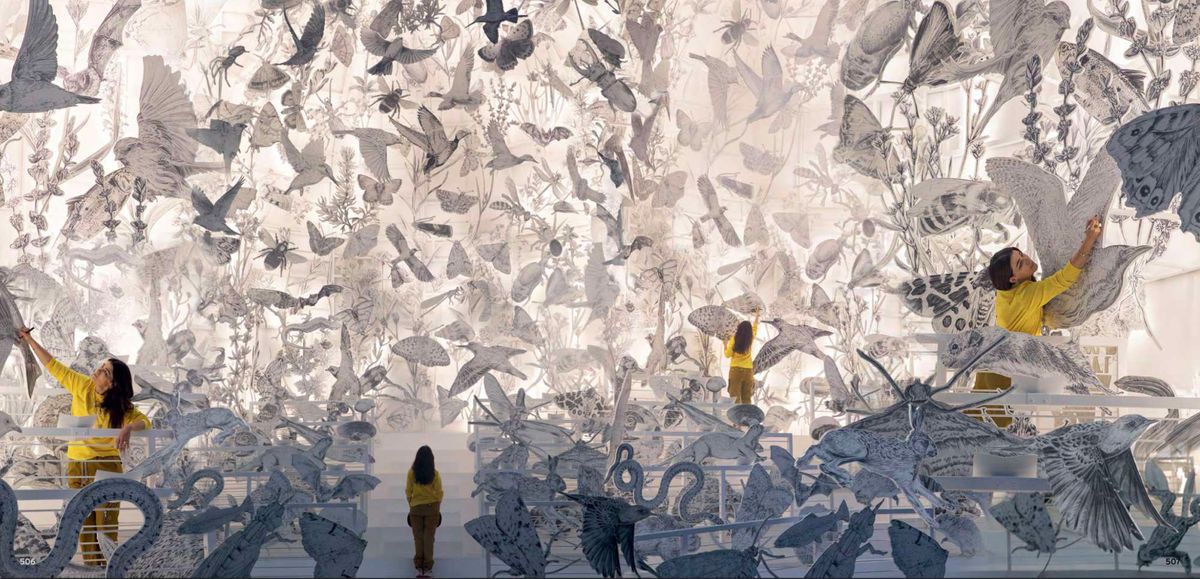 Es Devlin installation featuring hand-drawn birds, insects, and botanical illustrations suspended in a luminous space, with Es Devlin in yellow arranging the artwork.