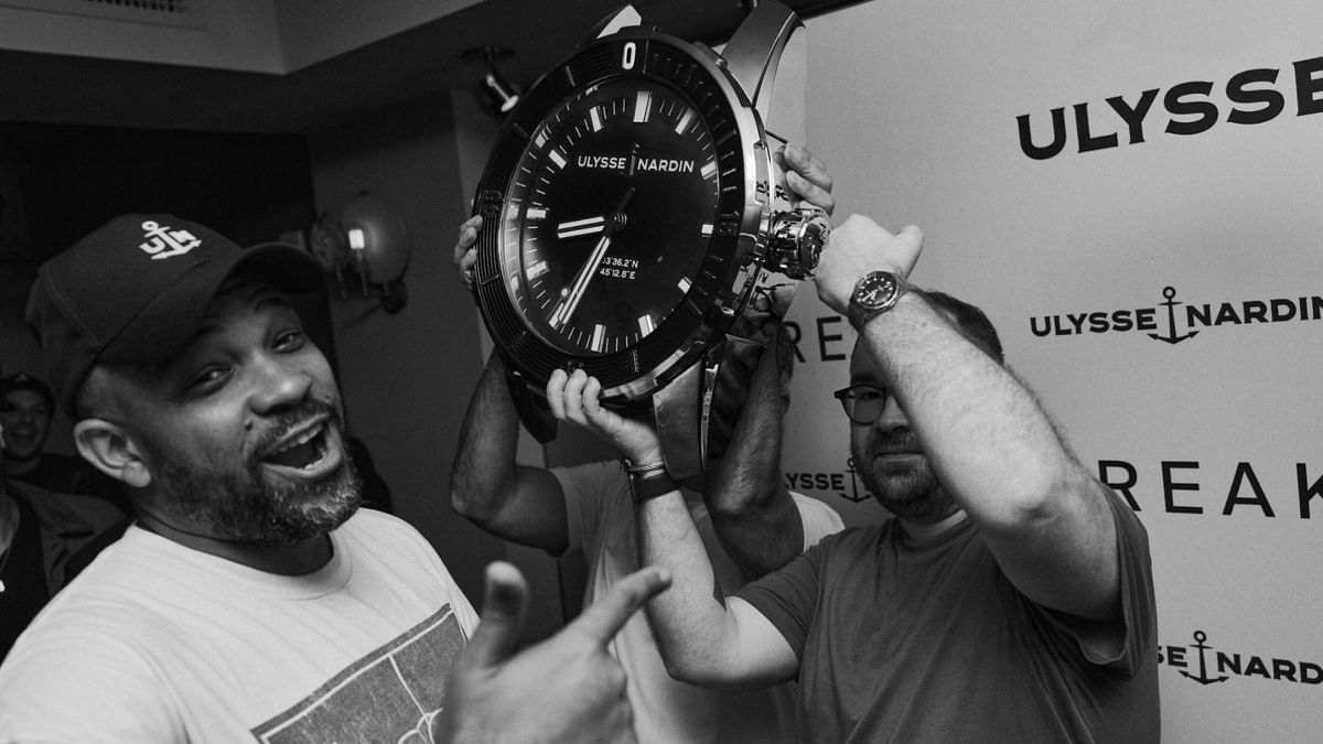 Perri Dash holding a large Ulysse Nardin wall clock during New York Watch Week, smiling in conversation with attendees.