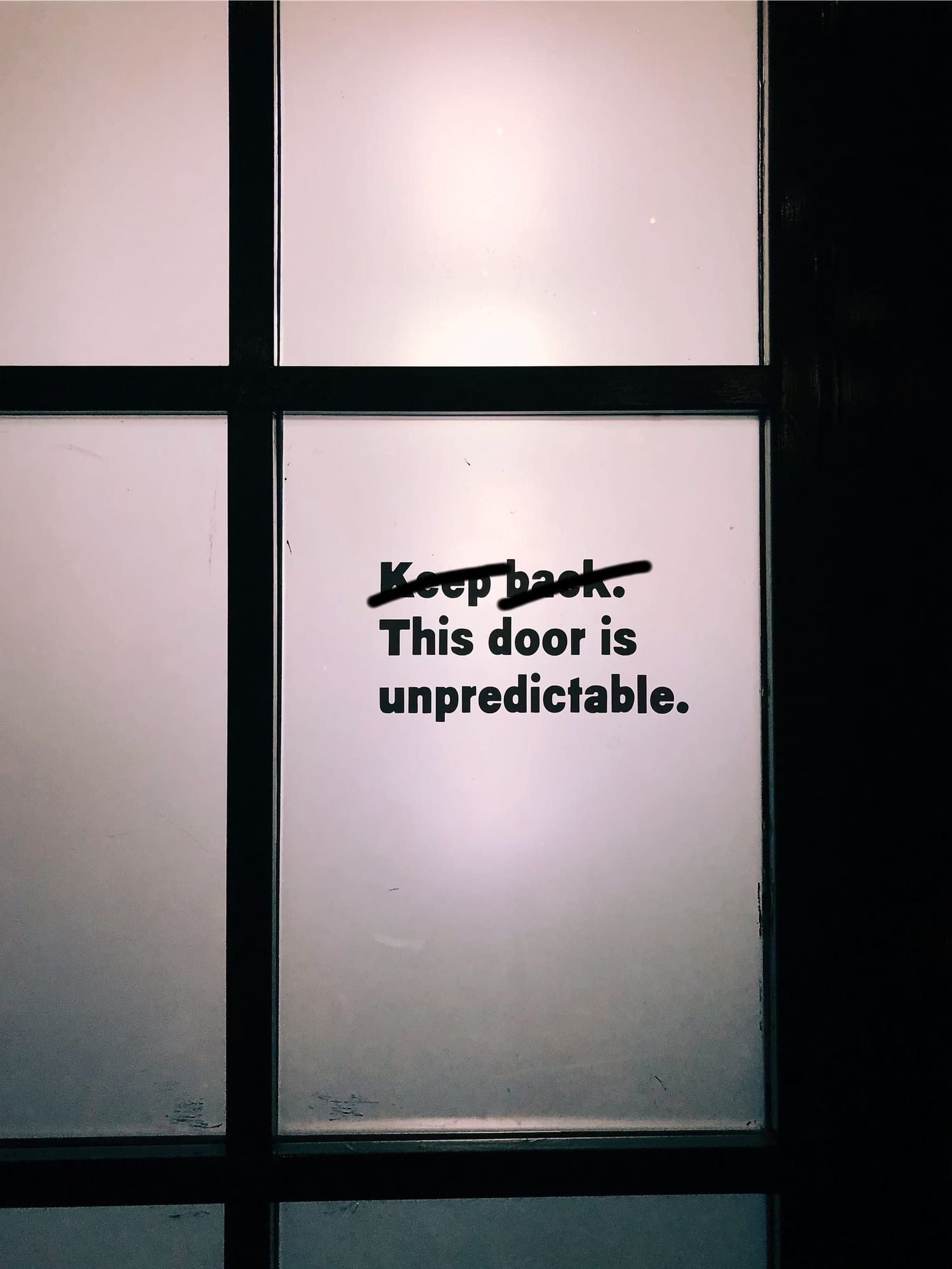 A photo of text on a door—"Keep back. This door is unpredictable.—with "keep back" crossed out.