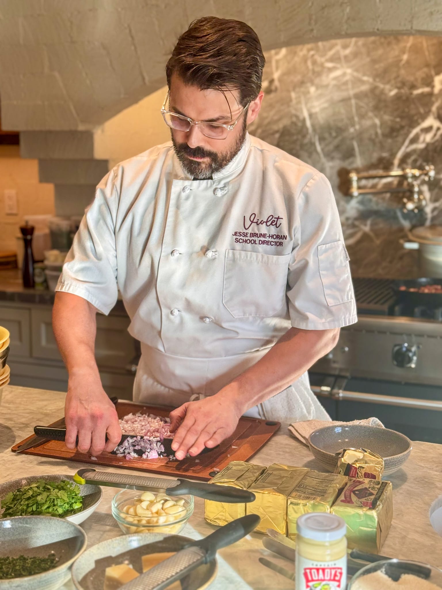 Chef Jesse Brune at Violet Cooking School mincing shallots surrounded by ingredients. He's wearing a white chef's jacket and apron.