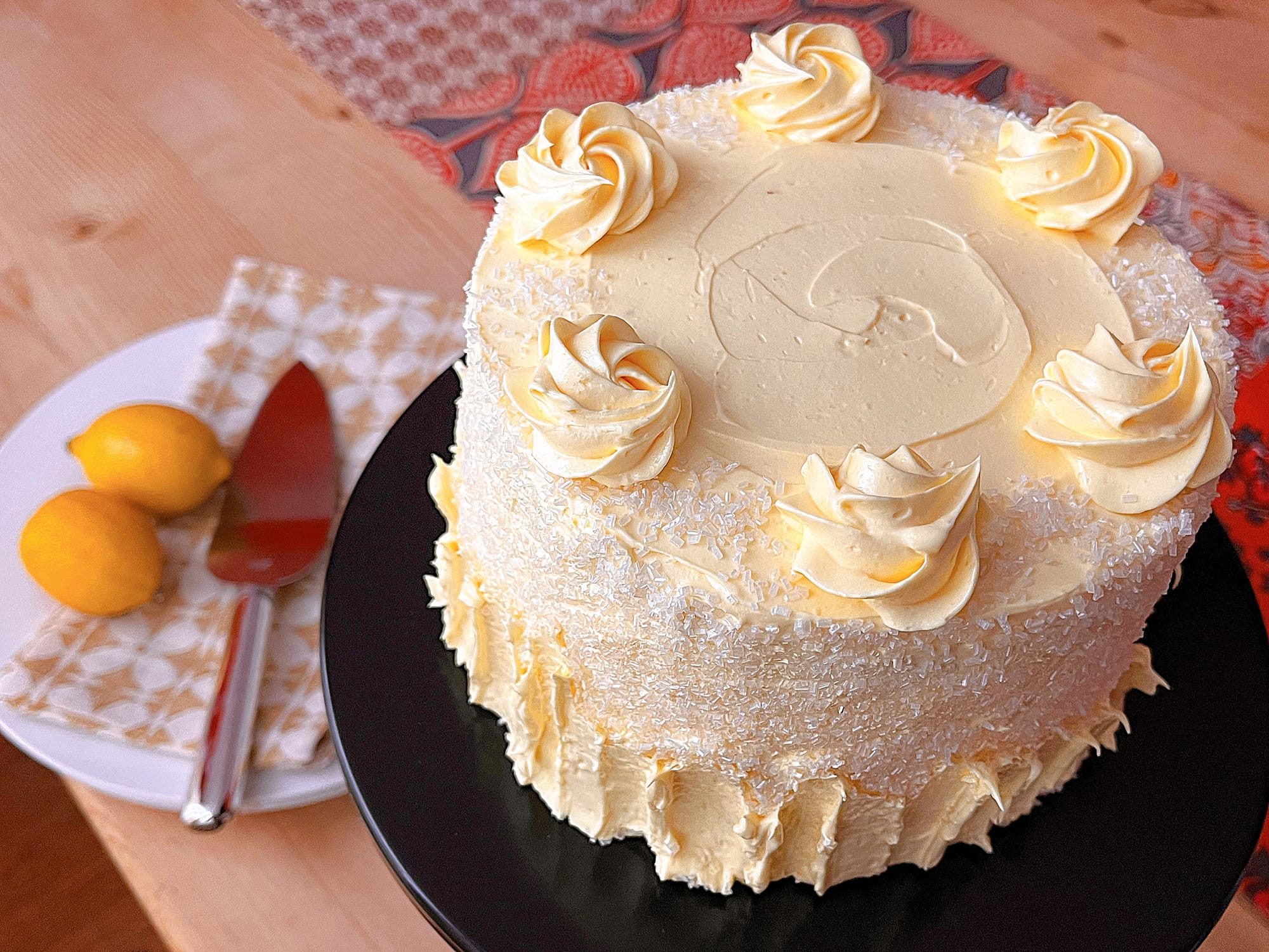 Lemon layered cake with Italian buttercream on a wooden surface next to a plate with a cake knife, linen napkin, and some bright yellow lemons for decor.