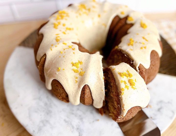 Orange and cardamom Bundt cake with a slice being lifted out, on a marble plate against a white tile backdrop
