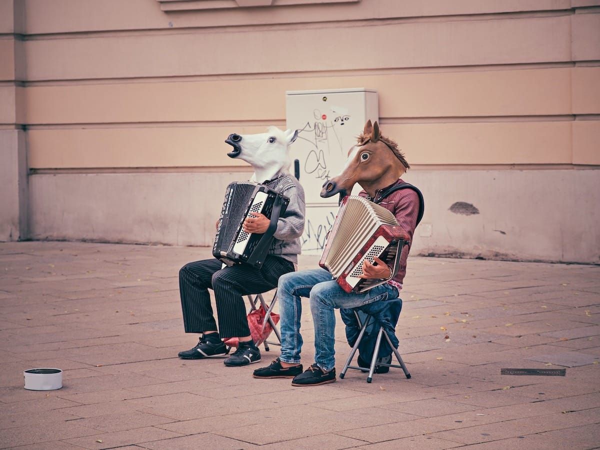 two person wearing horse heads sitting on folding chairs while playing accordions beside brown concrete building