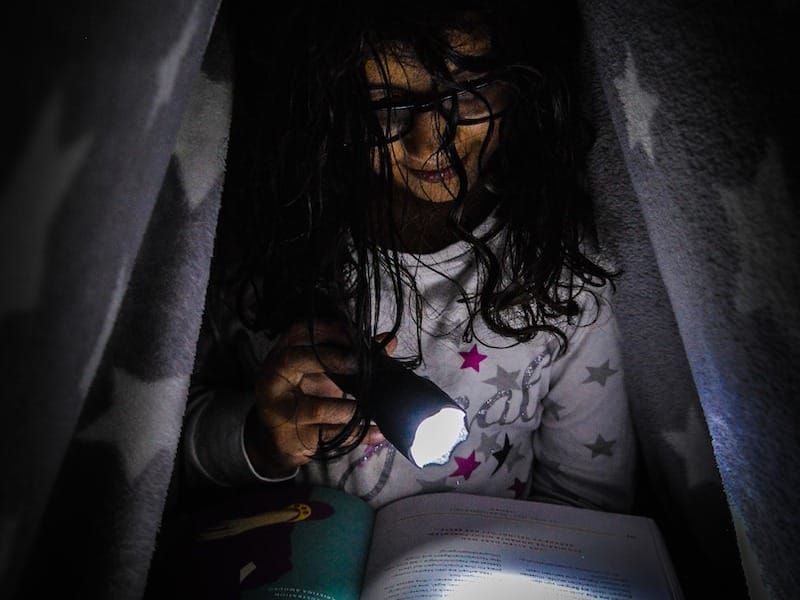 girl under the blanket reading by flashlight