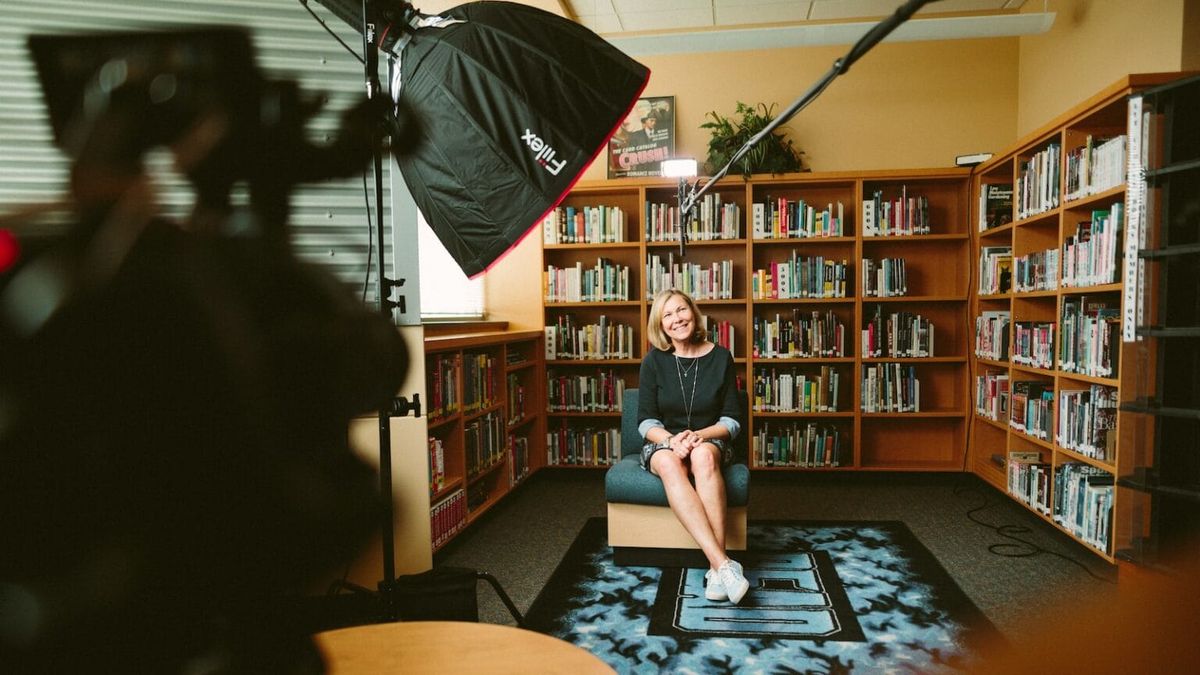 Woman behind a film camera sitting between three bookshelves.