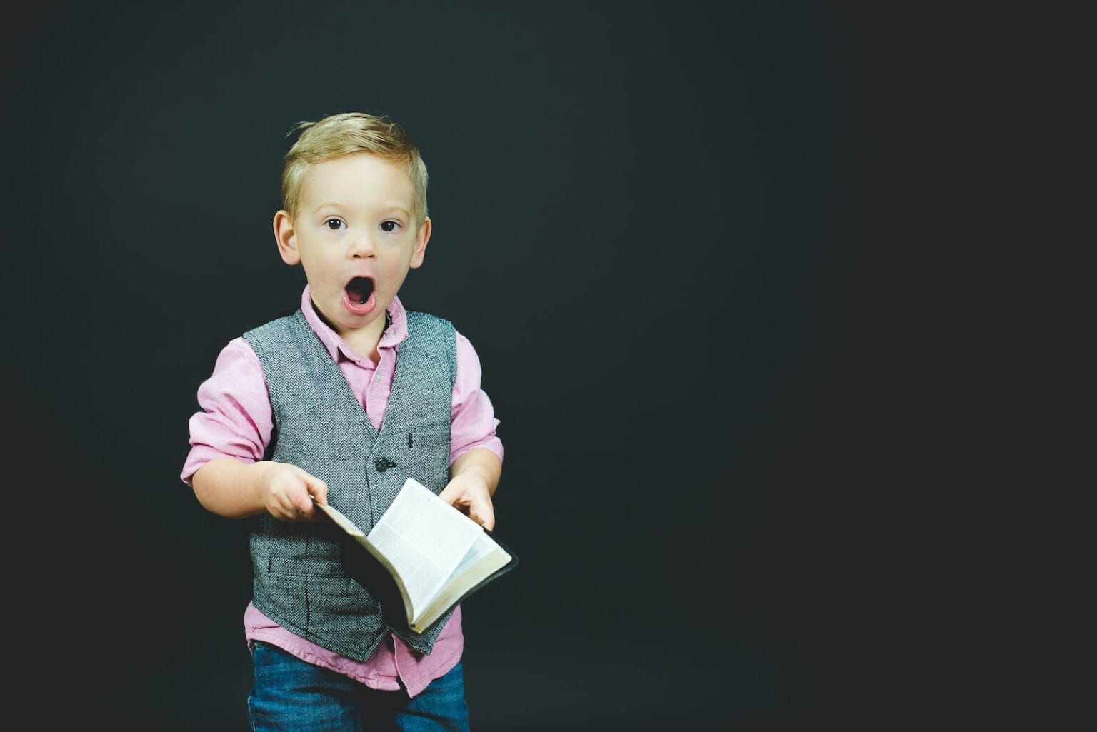 A boy in a grey vest mid-sentence from reading a book