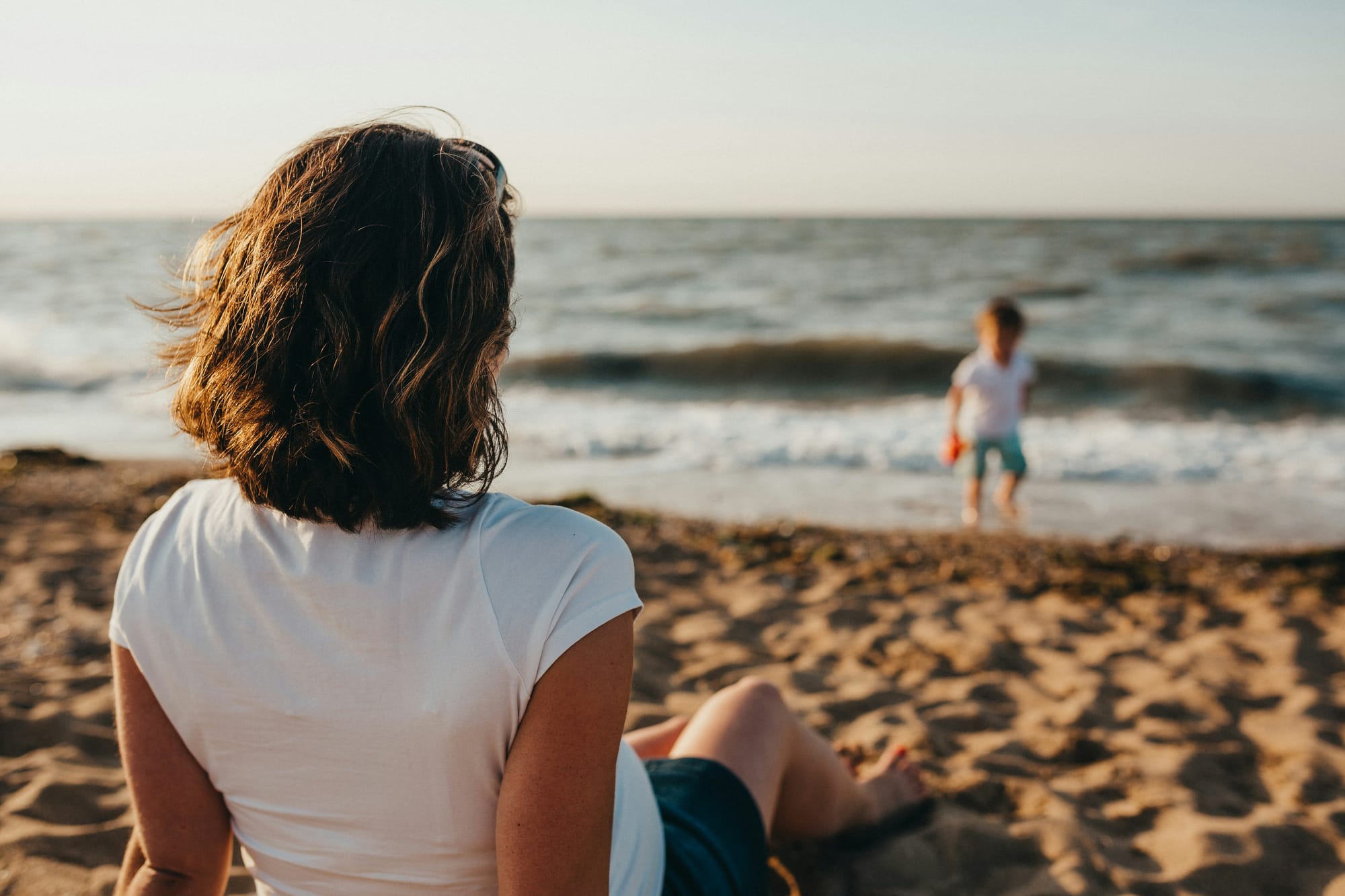 Photograph of a mother sitting on the beach and looking on as her son plays in the waves at the shoreline