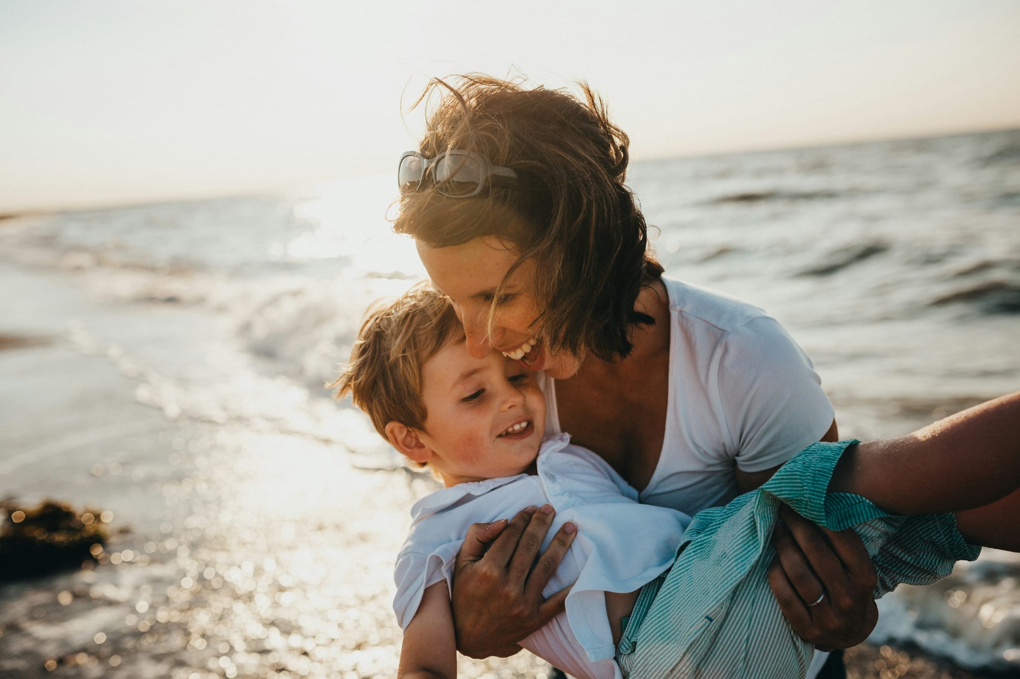 Photograph of a mother and son at the beach with the mother holding her son over the waves at the shoreline