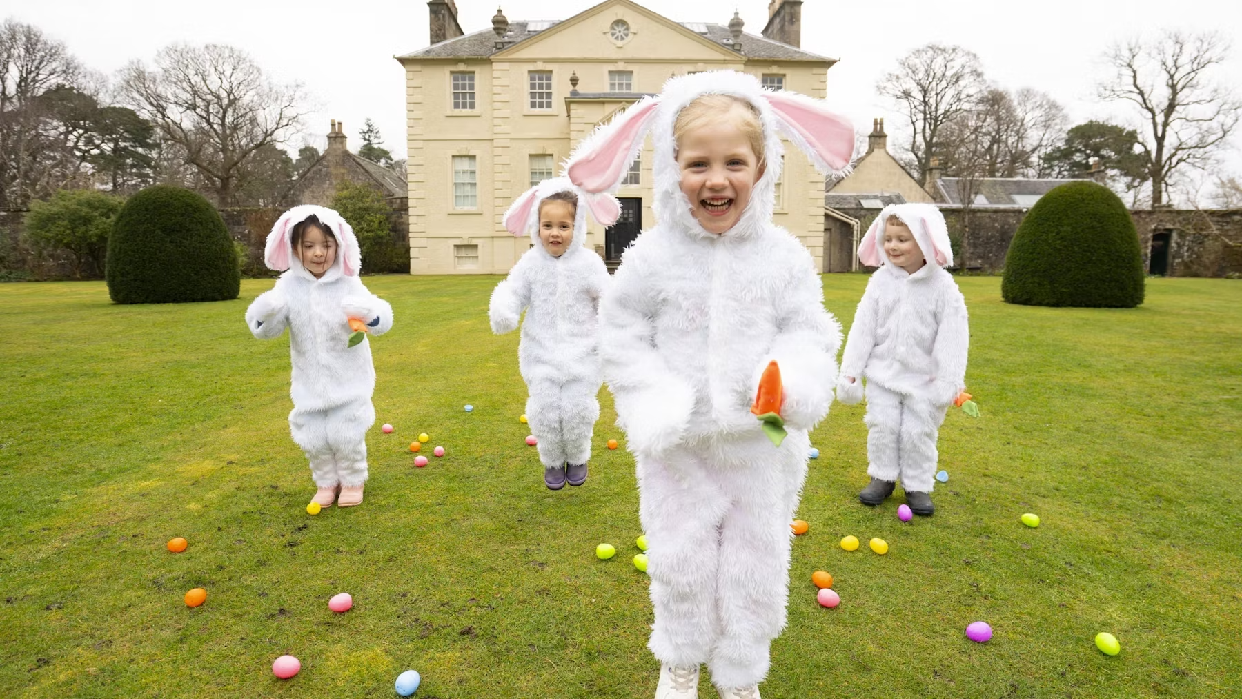 children in rabbit costumes play on a lawn in front of a stately home 