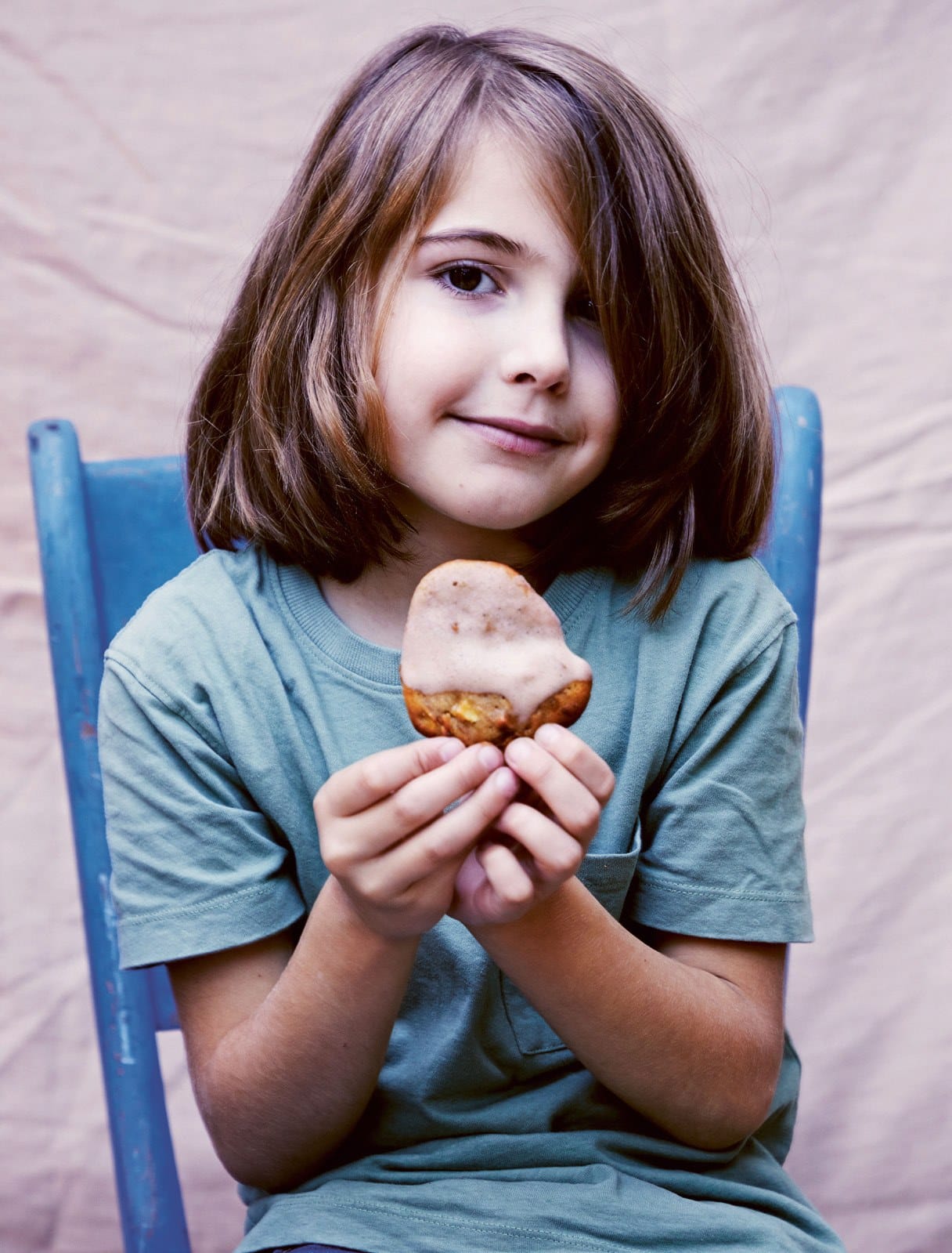 A smiling child on a blue chair holding a soft apple and cinnamon cookie topped with pink icing
