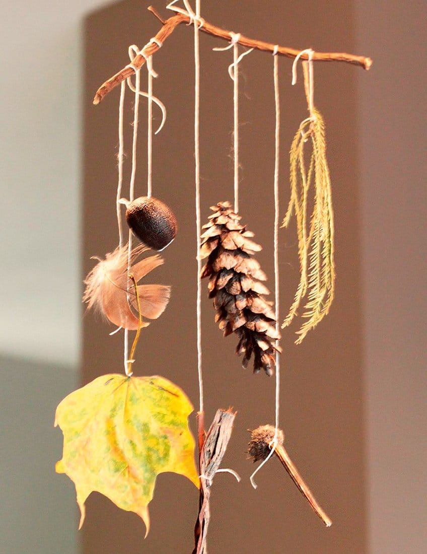 A nature mobile made from a stick hung with a pine cone, feather, acorn and autumn leaf